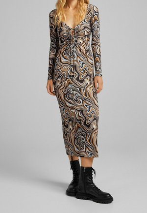 Frau trägt ein langärmeliges, Midi-Kleid mit einem braun, schwarz, beige und hellblau gewundenen Muster, kombiniert mit schwarzen Schnürstiefeln im Combat-Stil.