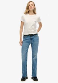 Camiseta de algodón blanca con un pequeño logotipo negro, combinada con jeans rectos azules y botas de tobillo de cuero negro. Atuendo simple y casual.