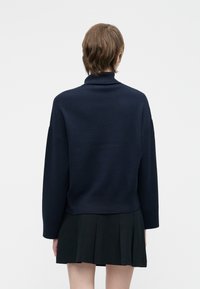 Temno modra pletena pulover s visokim ovratnikom, spušenimi rameni in rebrastimi manšetami, kombiniran s črno plisirano krilo.