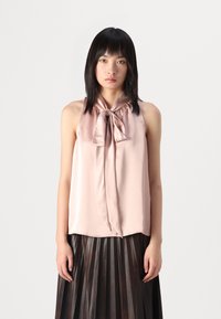 Top in satin rosa chiaro con dettaglio a fiocco frontale, design senza maniche e texture liscia. Indossato con una gonna plissettata scura, che mette in risalto il contrasto.