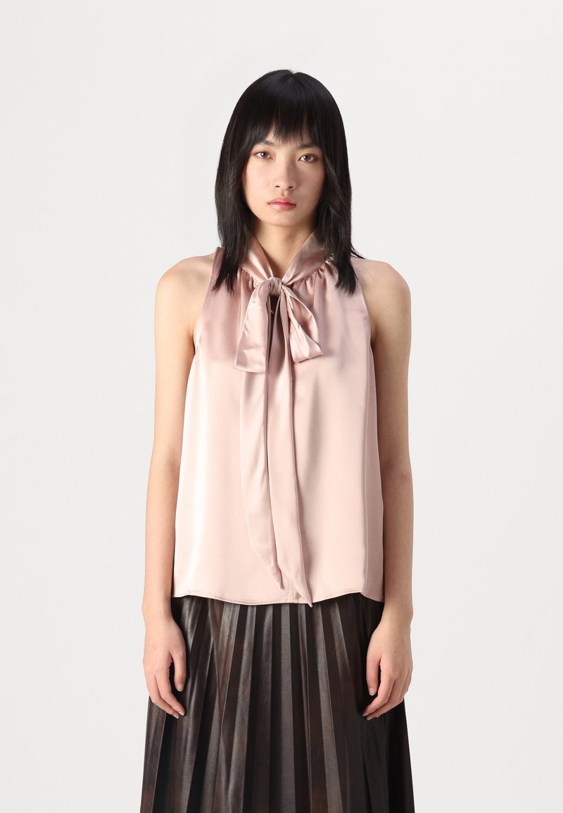 Top in satin rosa chiaro con dettaglio a fiocco frontale, design senza maniche e texture liscia. Indossato con una gonna plissettata scura, che mette in risalto il contrasto.
