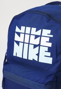 Tamno plavi ruksak s teksturiranom tkaninom, s velikim bijelim "NIKE" logotipom na prednjoj strani i zatvaranjem na patentni zatvarač na vrhu.