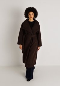 Abrigo largo marrón con textura y amplio cuello, con cinturón atado y dos bolsillos laterales. Llevado sobre una camiseta negra y combinado con pantalones oscuros acampanados.