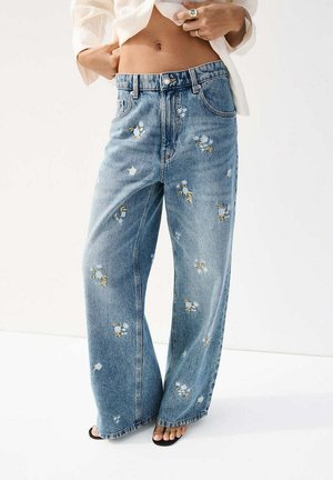 Jean denim bleu à jambes larges avec une petite broderie florale blanche et jaune, porté avec des sandales noires et un chemisier court et léger.