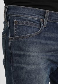 Vaqueros de mezclilla azul oscuro con un diseño clásico de cinco bolsillos, costuras naranjas contrastantes y una sutil textura desgastada.