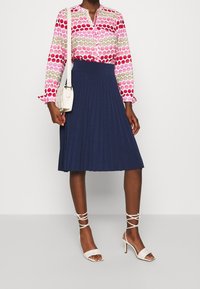 Långärmad blus med rosa och beige cirkulärt mönster, tillsammans med en navblå plisserad kjol. Vita klackar med remmar och en liten vit axelväska.