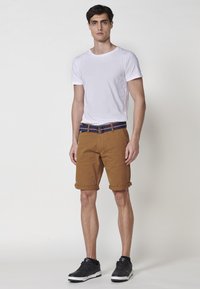 Beige Baumwollshorts mit umgeschlagenen Säumen, ausgestattet mit einem blauen Gürtel mit roten Akzenten. Getragen zu einem schlichten weißen T-Shirt mit kurzen Ärmeln und schwarzen Sneakers.