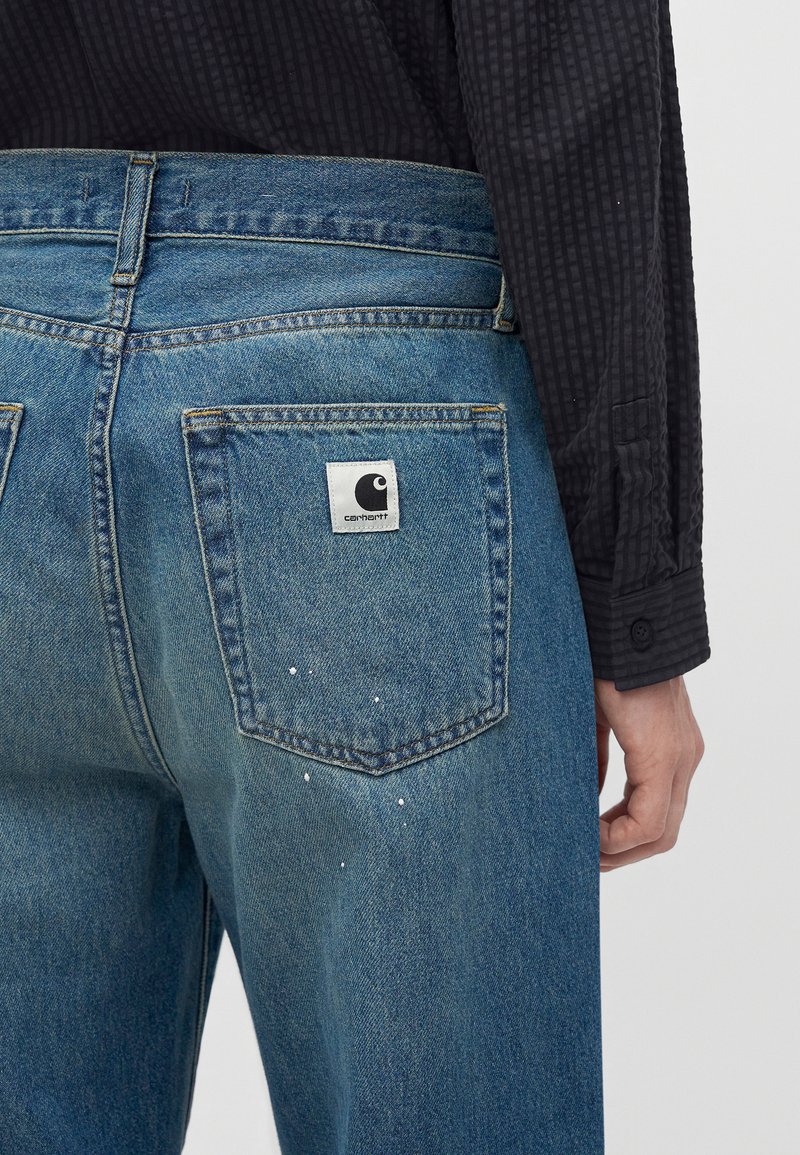 Vista posteriore di una persona che indossa jeans Carhartt blu con macchie di vernice bianca e una camicia nera a maniche lunghe e texture.