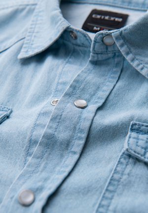 Jasnoniebieska koszula denimowa z kołnierzykiem, zapięciem na zatrzaski i dwoma kieszeniami na piersi, o miękkiej fakturze i widocznych szwach.