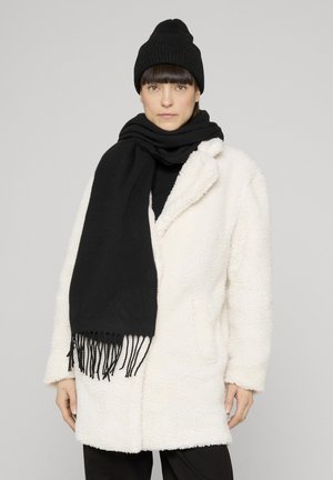 Frau trägt einen weißen, flauschigen Mantel, eine schwarze Strickmütze und einen langen schwarzen Schal und steht vor einem schlichten, hellen Hintergrund.