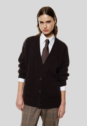 Cardigan marrone scuro realizzato in materiale morbido, con scollo a V, chiusura con bottoni e tasche. Abbinato a una camicia bianca e una cravatta marrone.