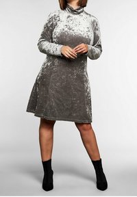 Žena v dlouhé stříbrné sametové šatech s dlouhými rukávy a černými kotníkovými botami, stojící s volně spojenýma rukama.