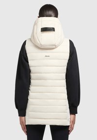 Puffweste in Creme mit einem gesteppten Design, einer großen Kapuze und einem schwarzen Logo auf der Kapuze. Kombiniert mit einem schwarzen Langarmshirt.