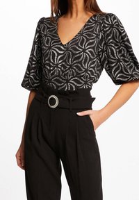 Svart topp med silvervirvlar och v-ringning, tillsammans med högmidjade svarta byxor med veck och en dekorativ bältesspänne.