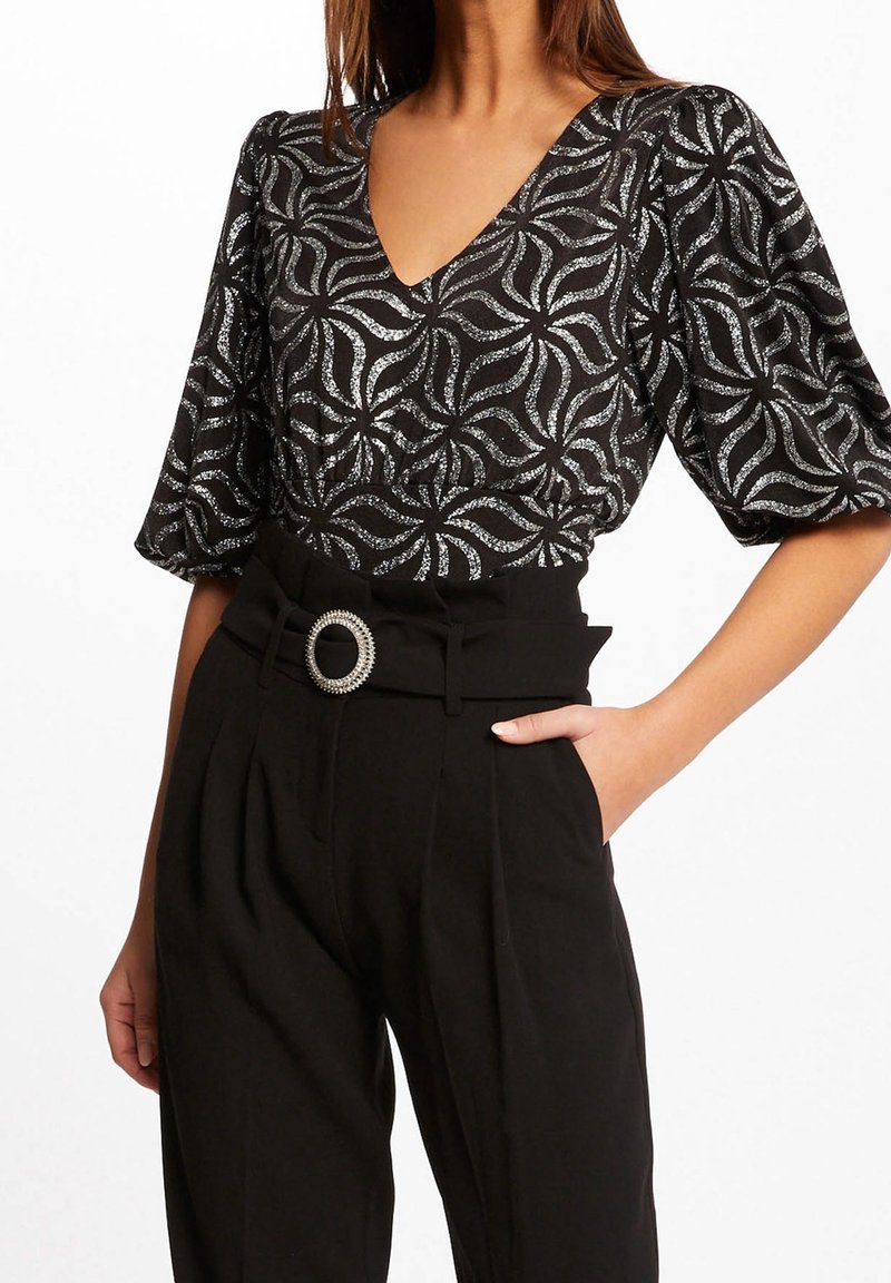 Svart topp med silvervirvlar och v-ringning, tillsammans med högmidjade svarta byxor med veck och en dekorativ bältesspänne.