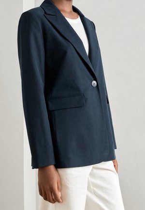 Person iført en enkelt-knappet navy blazer over en hvid top og hvide bukser, stående mod en ensfarvet lys baggrund.