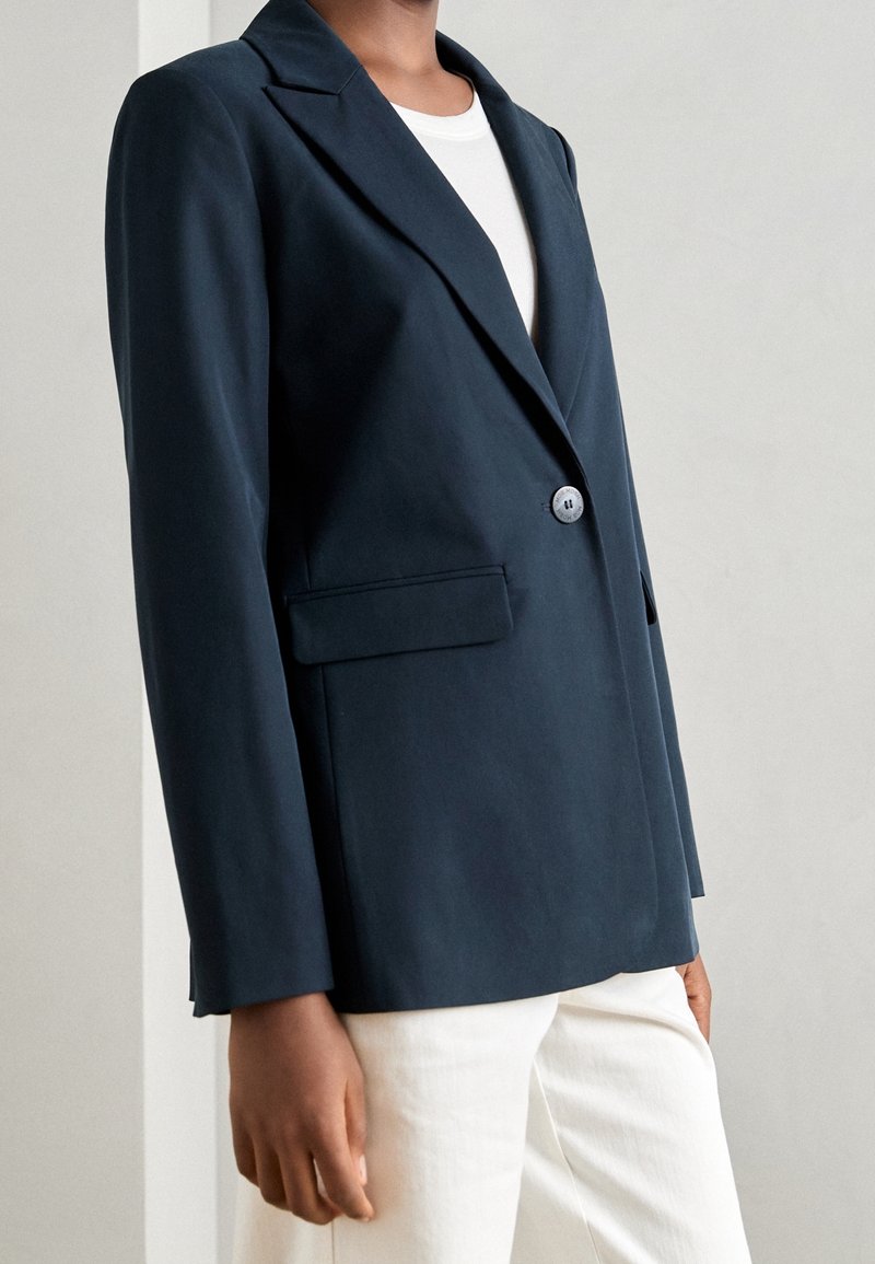 Personne portant un blazer bleu marine à bouton unique sur un haut blanc et un pantalon blanc, debout devant un fond clair uni.