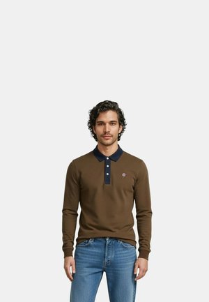 Homme debout portant un polo à manches longues marron avec un col bleu marine et un jean bleu, sur un fond blanc uni.