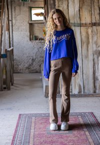 Blauwe crop sweatshirt met de tekst "Queens", gecombineerd met bruine flared broeken. Witte sneakers maken de outfit af. Natuurlijke binnenomgeving.