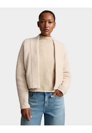 Donna che indossa un cardigan aperto a coste color crema sopra un maglione a collo alto crema e jeans azzurri chiari, in piedi con una mano in tasca.