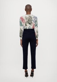 Sveter s dlhými rukávmi v krémovej farbe s farebnými vzormi stromov, v kombinácii s prispôsobenými nohavicami tmavomodrej farby a čiernymi lodičkami, z pohľadu zozadu.