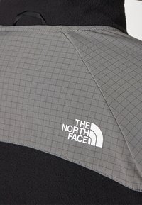 Primo piano di una giacca in pile nera con pannello a motivi grigi sulla spalla, caratterizzata dal logo The North Face in bianco.