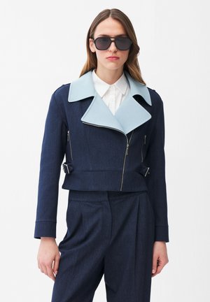 Kobieta ubrana w ciemnoniebieski garnitur z szerokim, jasnoniebieskim kołnierzem, białą koszulę z kołnierzykiem oraz duże czarne okulary przeciwsłoneczne, stojąca na białym tle.