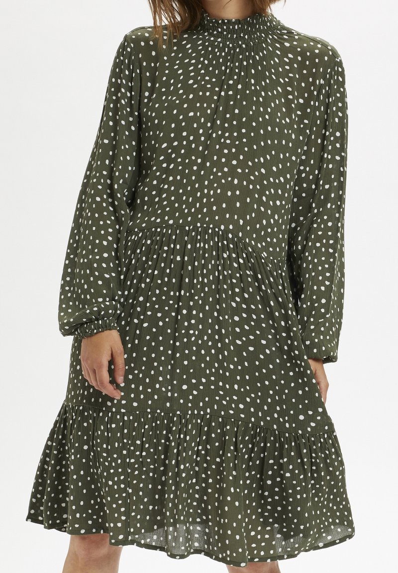 Robe verte mi-longue avec des pois blancs irréguliers, des longues manches froncées et un ourlet volanté portée par une personne.