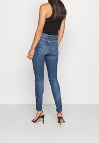 Jeans ajustados de mezclilla azul oscuro, con cintura alta, bolsillos traseros y un dobladillo ligeramente deshilachado. Combinados con tacones negros.