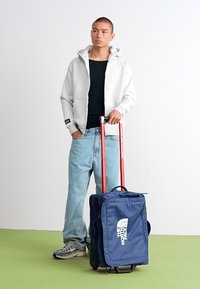 Valise à roulettes en tissu bleu marine avec un logo blanc, poignées rétractables rouges et roues robustes. Portée avec un sweat à capuche gris décontracté et un jean clair.