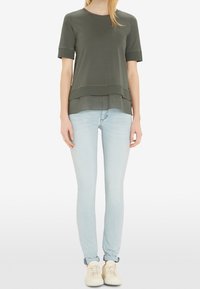 Olivengrøn kortærmet top med lagdelt kant, parret med lyseblå skinny jeans og hvide sneakers. Glat stoftekstur.