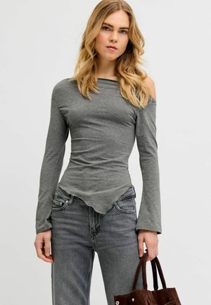 Žena s dlhými blond vlasmi oblečená v sivom top s dlhým rukávom a odhalenými ramenami a sivých džínsoch, drží hnedú semišovú kabelku po boku.