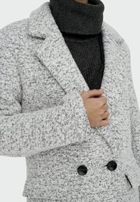 Persoon draagt een getextureerde grijs-witte blazer met dubbele knopen over een donkergrijze geribbelde coltrui, en houdt de revers vast.