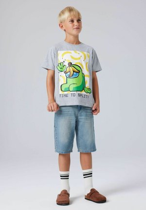 Garçon blond debout portant un t-shirt gris avec un personnage vert et un motif banane, un short en jean, des chaussettes blanches à rayures noires et des chaussons marron.