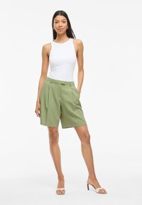 Shorts verdes ajustados con pliegues delanteros, cintura alta y lazos para cinturón, combinados con una camiseta sin mangas blanca ceñida y tacones en tonos neutros.