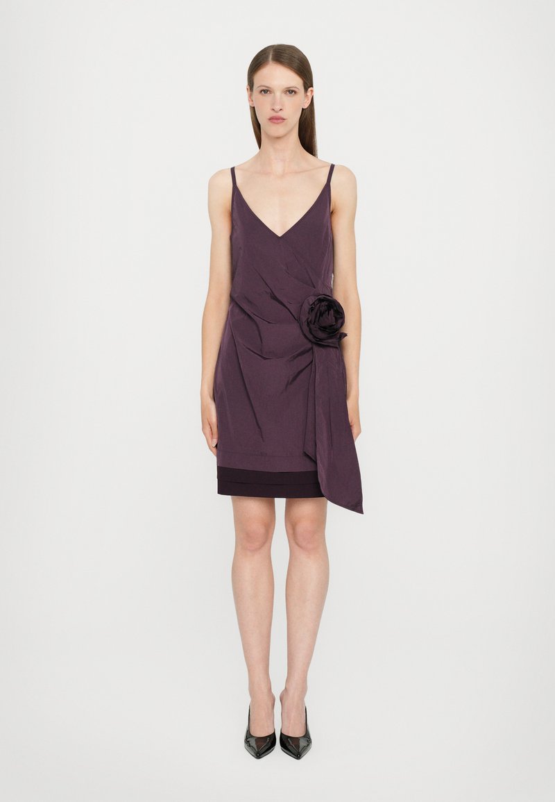 Purple short dress with a V-neck, adjustable straps, and a floral fabric accent. Smooth texture with a layered skirt. Black high-heeled shoes.