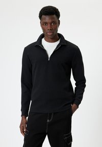Zwarte longsleeve pullover met een kraag en halve rits, met een textuur en geribbeld ontwerp. Draag het over een wit T-shirt, in combinatie met zwarte broek.