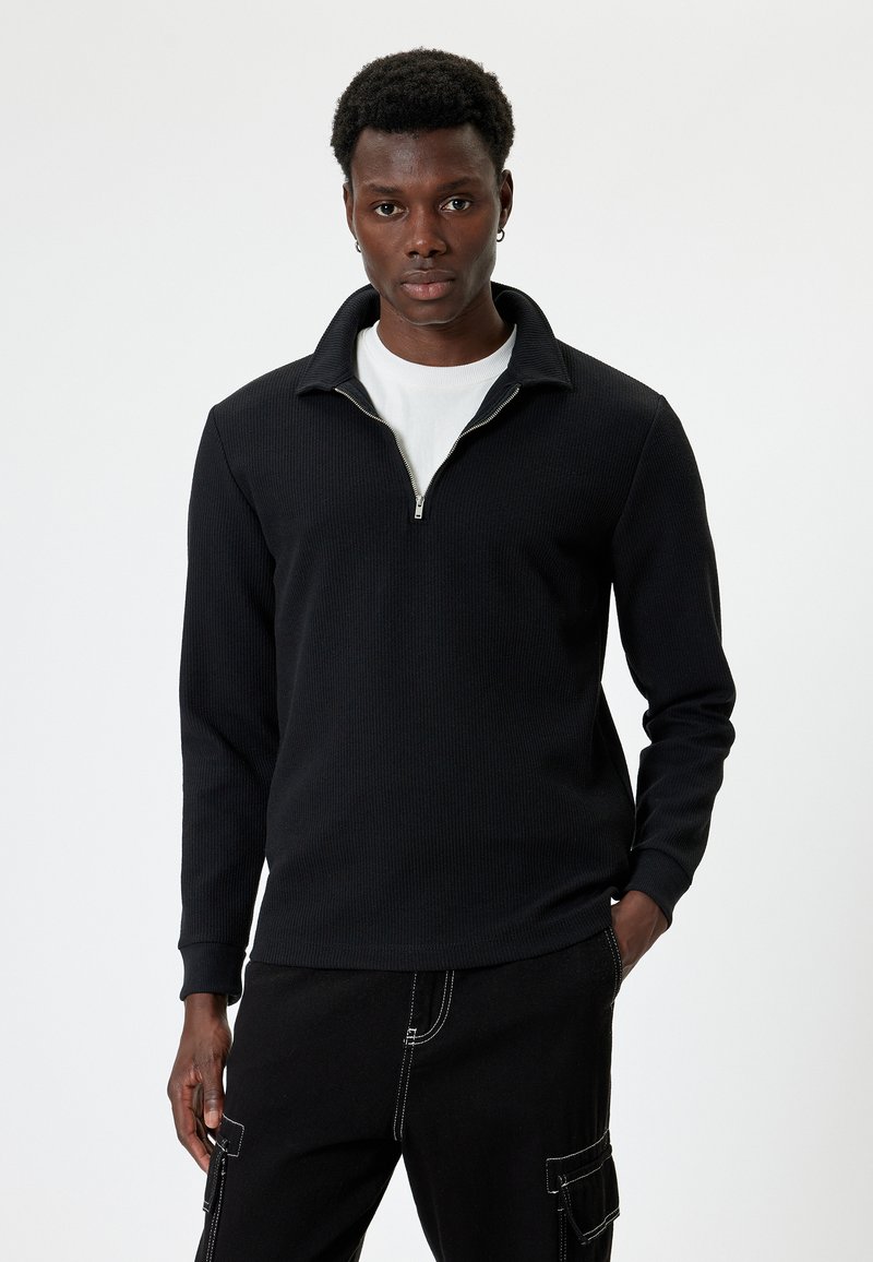 Zwarte longsleeve pullover met een kraag en halve rits, met een textuur en geribbeld ontwerp. Draag het over een wit T-shirt, in combinatie met zwarte broek.