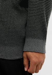 Pull en tricot texturé gris avec des bords côtelés aux poignets et à l'ourlet, main visible, démontrant un gros plan sur le tissage du tissu et la variation de couleur.