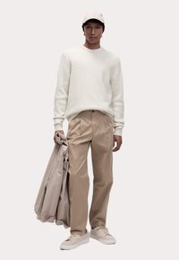 Weißer, strukturierter Pullover, beigefarbene, plissierte Hose, hält eine hellbeige Jacke. Kombiniert mit hellen Sneakern, wirkt das Gesamtbild neutralfarben.