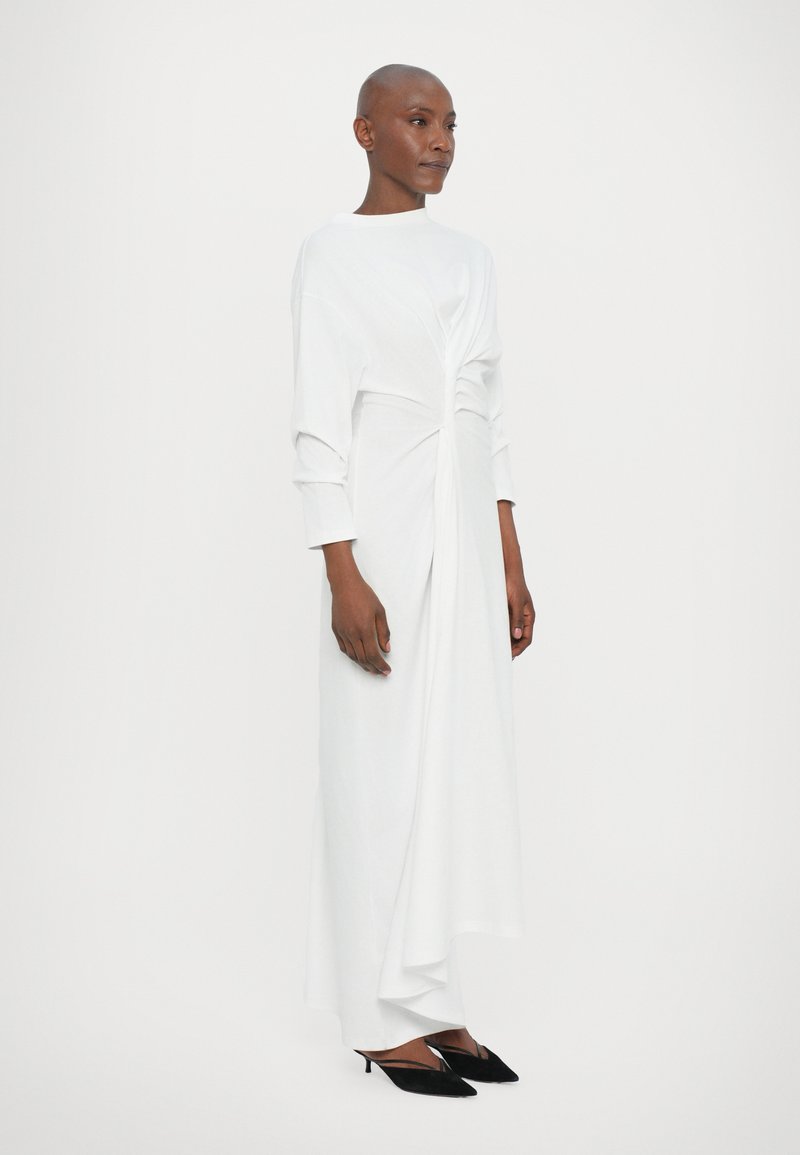Lang witte jurk van zacht materiaal, met een gedraaide voorzijde en lange mouwen, gecombineerd met zwarte pumps met een puntige neus.