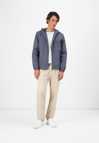 Veste légère bleu marine avec capuche, poignets élastiques et logo jaune. Portée par-dessus une chemise blanche et un pantalon beige, associée à des chaussures blanches.
