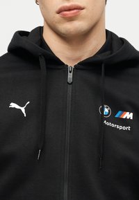 Κοντινό πλάνο ενός ατόμου που φοράει μαύρο φούτερ με φερμουάρ και τα λογότυπα Puma και BMW Motorsport στο στήθος.