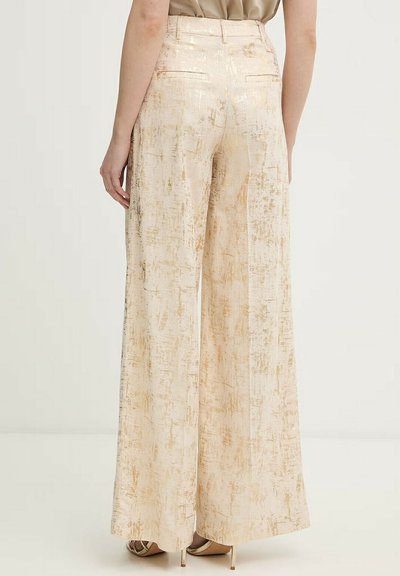 Vrouw draagt hoge taille, wijde crèmebroek met gouden textuurpatroon, gecombineerd met een beige blouse en beige sandalen met hoge hak.