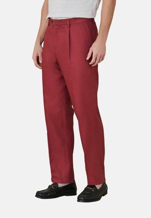 Mann trägt rote maßgeschneiderte Hose, schwarze Loafers und ein weißes gestreiftes Hemd, steht vor einem schlichten weißen Hintergrund.