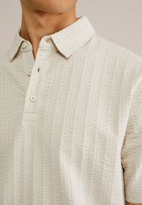 Polo shirt bej deschis cu un model texturat, guler cu nasturi și două nasturi la placket. Mâneci scurte și croială lejeră.