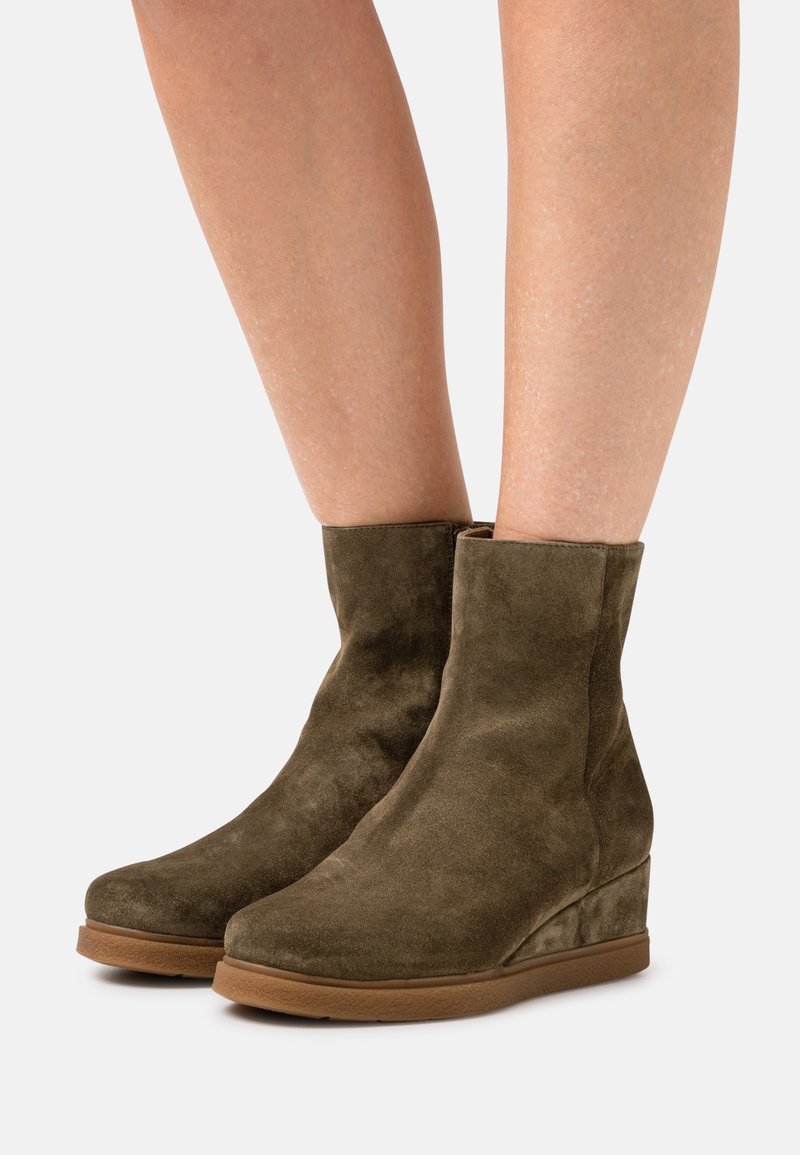 Bottines en daim marron à talons compensés portées sur des jambes nues sur un fond clair uni.
