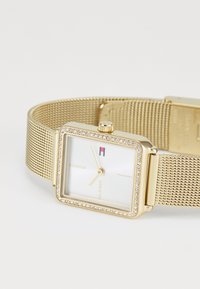 Montre-bracelet avec un bracelet en maille dorée, un boîtier rectangulaire, une lunette sertie de cristaux, des aiguilles dorées minimalistes et un petit logo rouge, blanc et bleu sur le cadran blanc.
