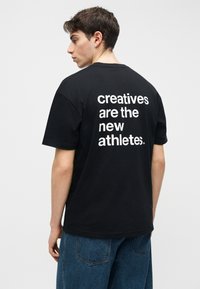 Noor mees lühikeste pruunide juustega, musta T-särgi ja siniste teksadega, T-särgil tekst: "loojad on uued sportlased."