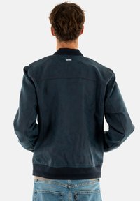 Chaqueta bombers de la marina en material texturizado, con cuello y puños acanalados. La vista trasera muestra un sutil detalle de costura y el brillo del tejido.